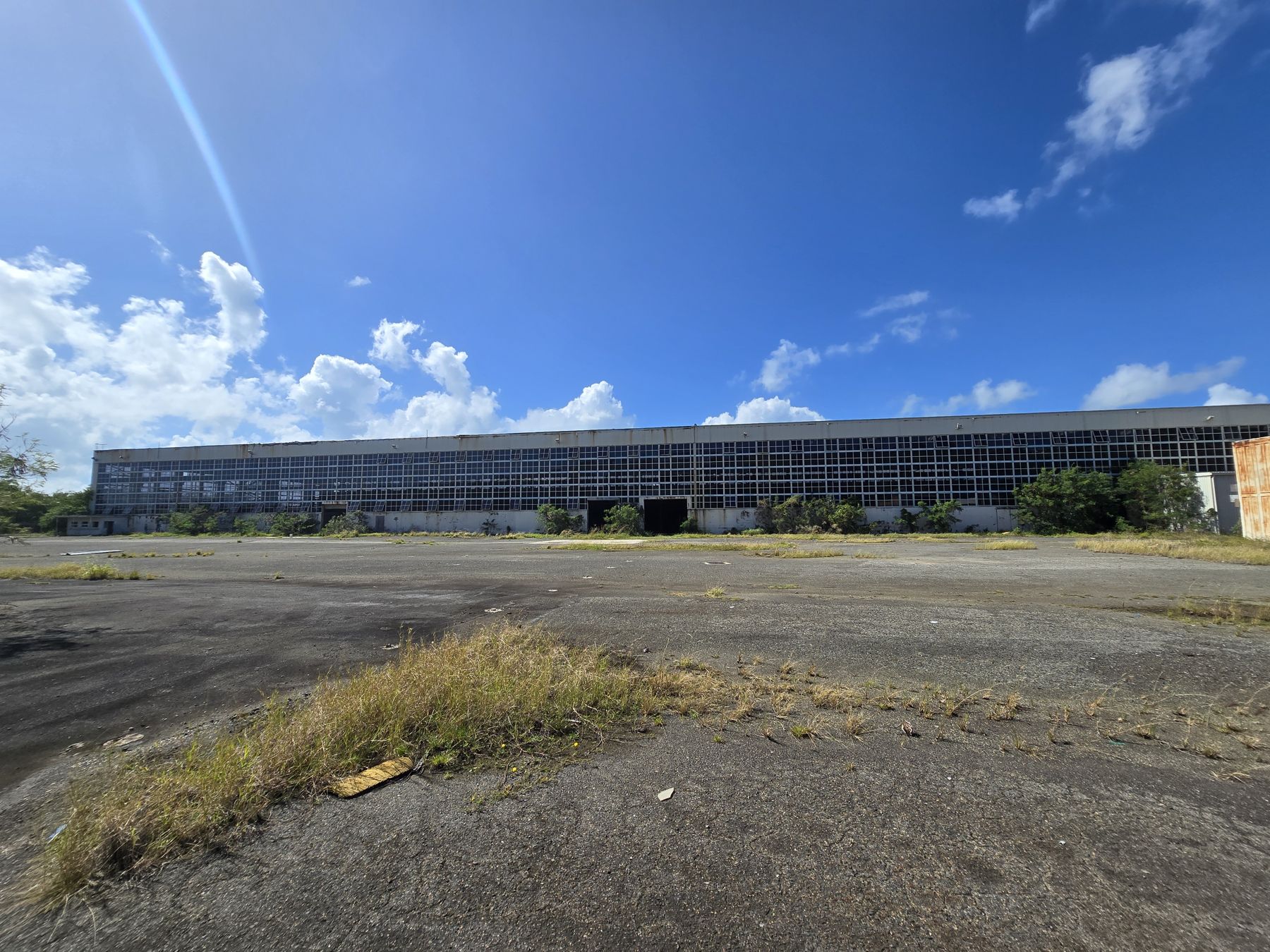 Fachada de vidrio de un hangar abandonado frente a una plataforma de asfalto con yerba